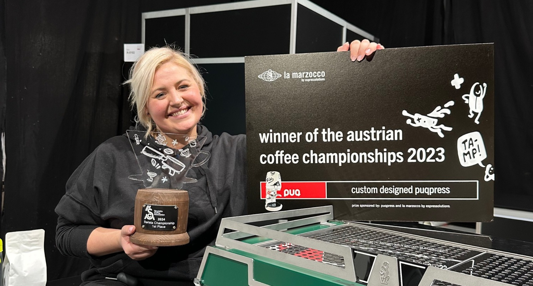 Barista Dominika Piotrowska sitting beside a green espresso machine and a sign proclaiming her the winner of the Austrian Barista Championships 2023